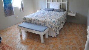 Een slaapkamer met een bed, nachtkastje, bankje en een raam met gordijnen.