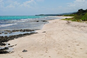 A sandy beach with footprints, a rocky shoreline, and a distant view of a green island under a clear sky. - 6-bedroom half house in Duncans Bay with AC, Security and manager on duty 24/7 (Duncans)