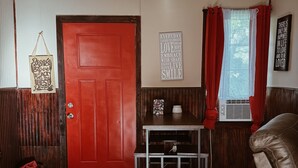 A room with a red door, a small table with a white coffee pot, a leather couch, and a window with red curtains.