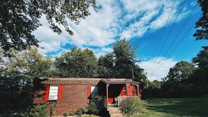A small, single-story wooden building with a porch and a red door, surrounded by a grassy yard and trees.