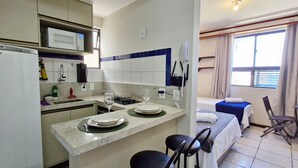 A compact hotel room with a kitchenette, a bed with a blue cover, a small dining area, and a window with curtains.