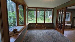 Quarto com janelas amplas, vista para uma floresta, tapete pequeno e um banco com um urso de pelúcia.