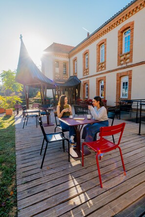 Un café al aire libre con mesas y sillas de madera, un espacio cubierto para sentarse y un edificio con paredes de ladrillo y ventanas arqueadas.
