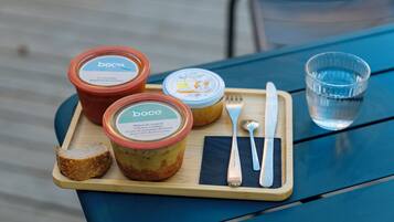 A wooden tray with jars of food, a piece of bread, cutlery, and a glass of water on a blue table.