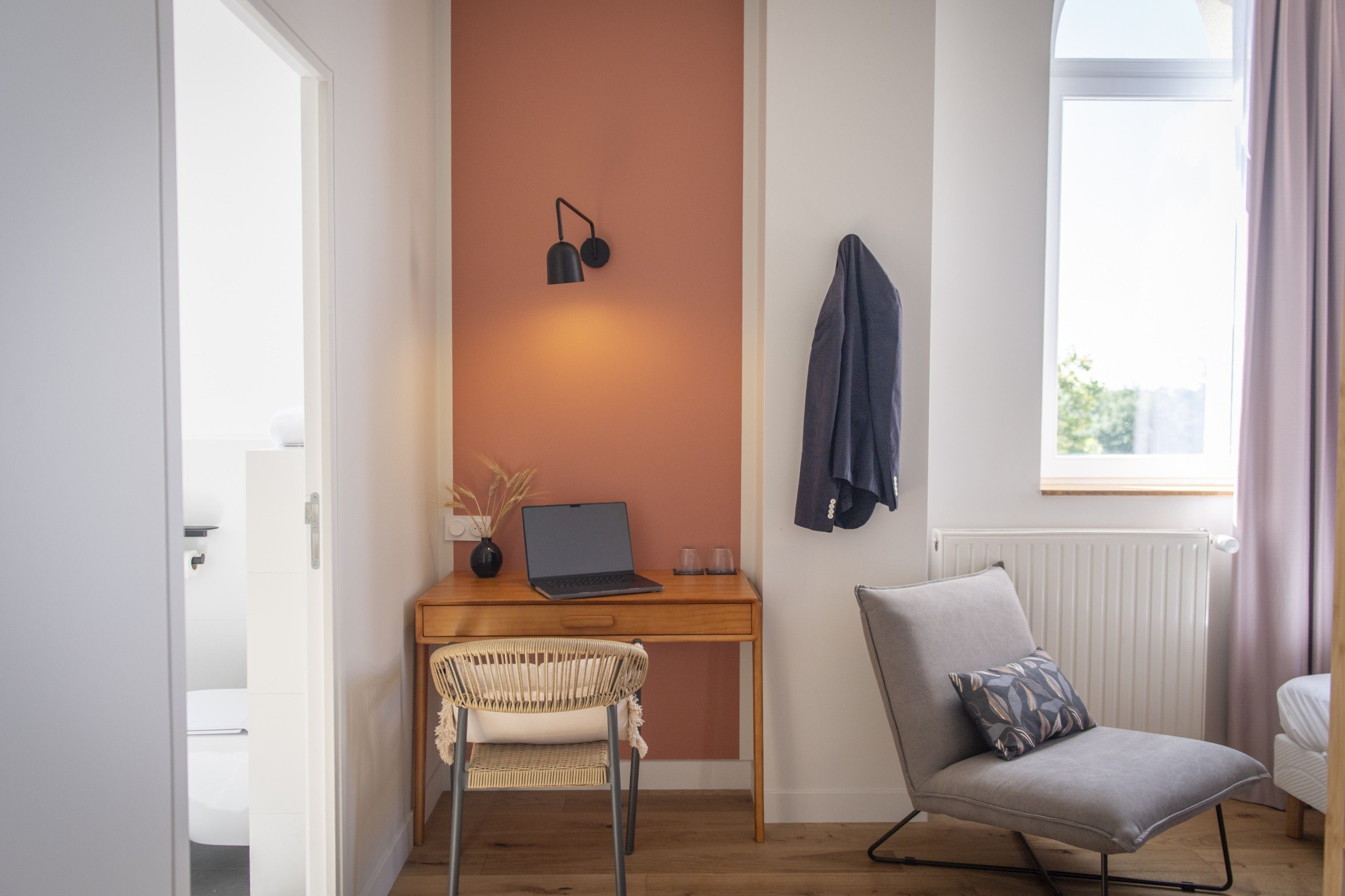 A modern room with a wooden desk, a laptop, a wicker chair, a grey armchair, and a window with curtains.