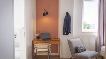 A modern room with a wooden desk, a laptop, a wicker chair, a grey armchair, and a window with curtains.