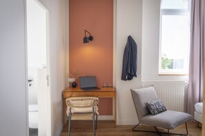 Una habitación moderna con un escritorio de madera, una computadora portátil, una silla de mimbre, un sillón gris y una ventana con cortinas.