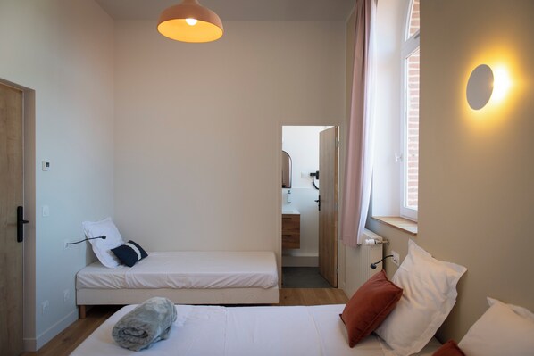 Un dormitorio pequeño y bien iluminado con dos camas, una ventana con persianas, una puerta y una lámpara.