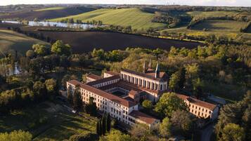 Vista aérea de un edificio grande e histórico con varias alas y un patio central, rodeado de campos verdes exuberantes y una masa de agua al fondo.
