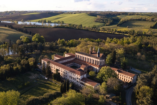 An aerial view of a large, historic building with multiple wings and a central courtyard, surrounded by lush green fields and a body of water in the background.