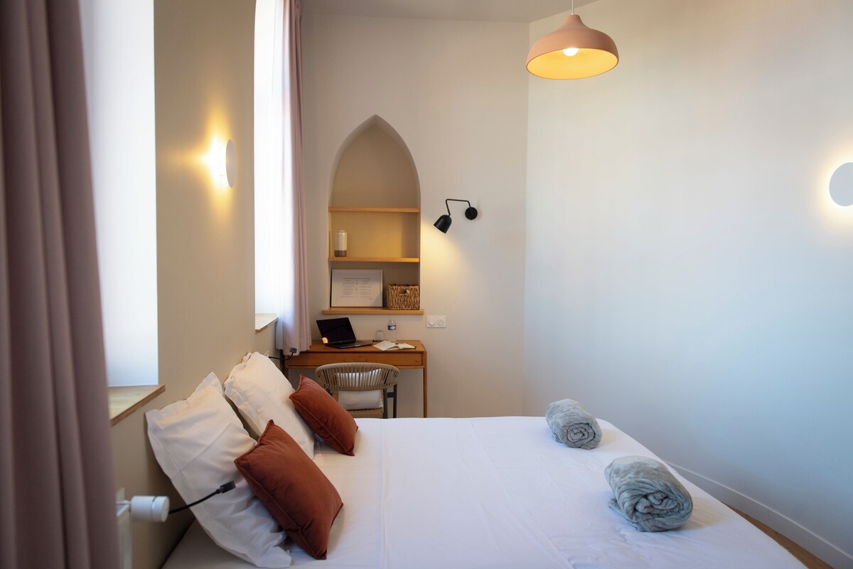 Une chambre à coucher avec un lit, des oreillers, un bureau avec un ordinateur portable, une chaise et une lampe.