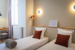 A hotel room with two beds, a desk, a chair, a radiator, a wall-mounted light, and a framed document on the wall. - Hotellerie De L'abbaye Du Désert (Thil)