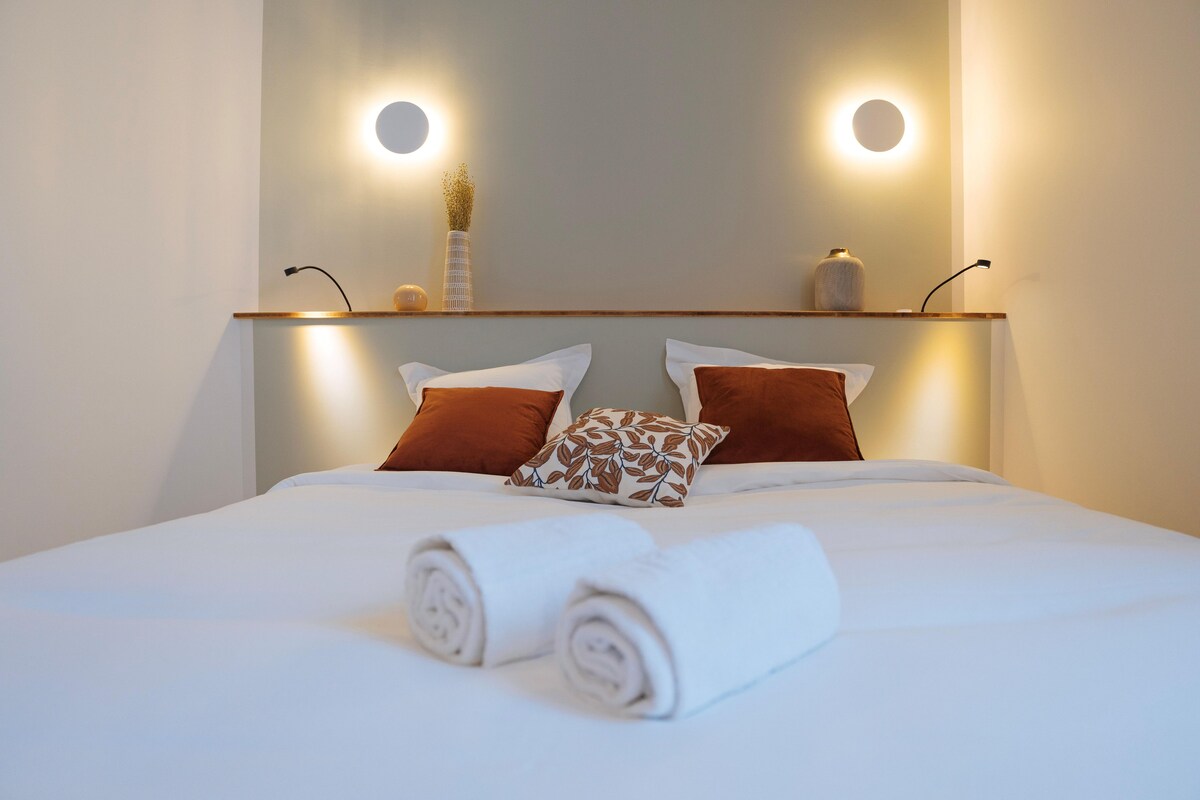 A neatly made bed with rolled towels and two pillows, a bedside table with a vase and a lamp, and wall-mounted circular lights.