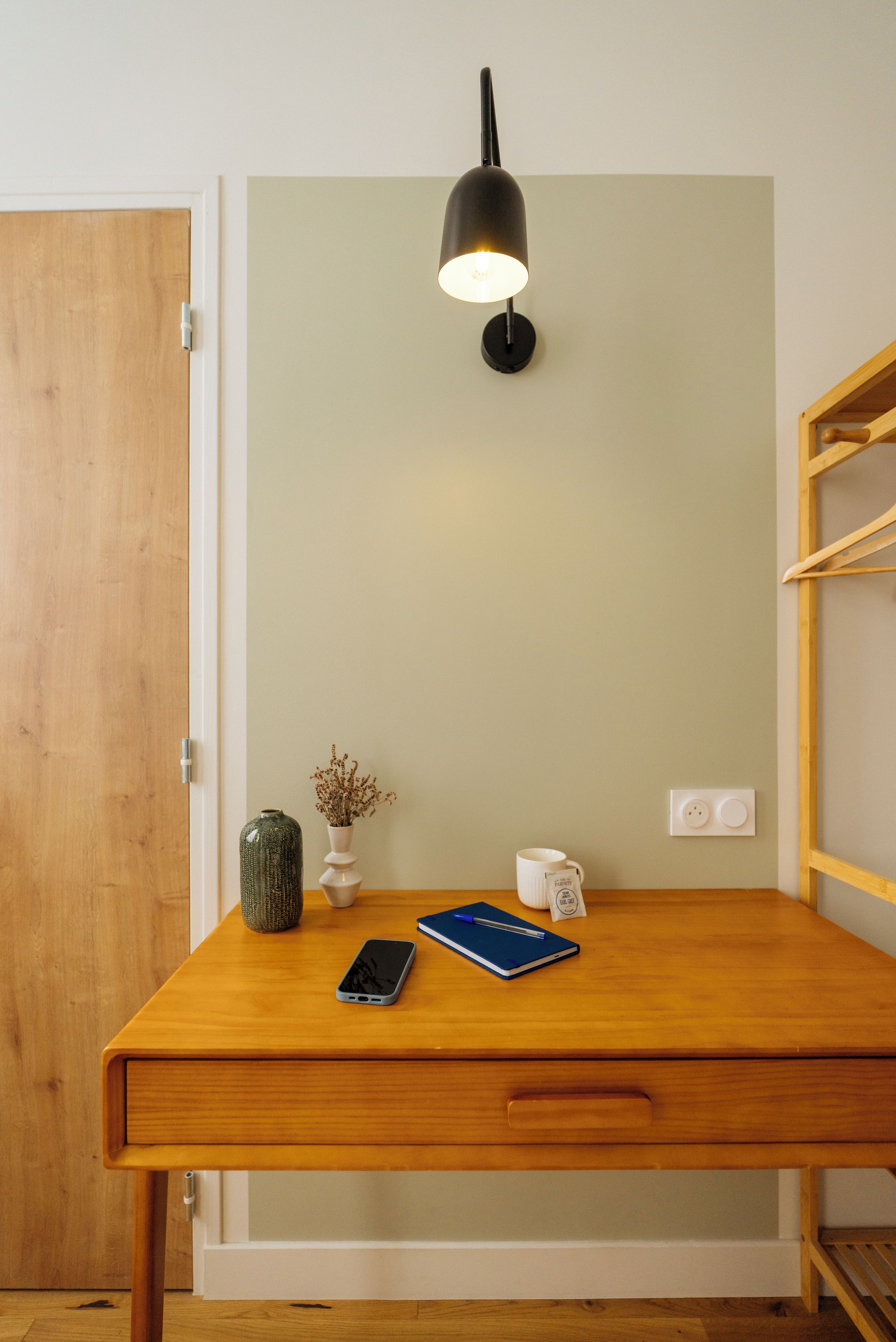 A wooden desk with a lamp, a vase, a mug, a notebook, and a smartphone.