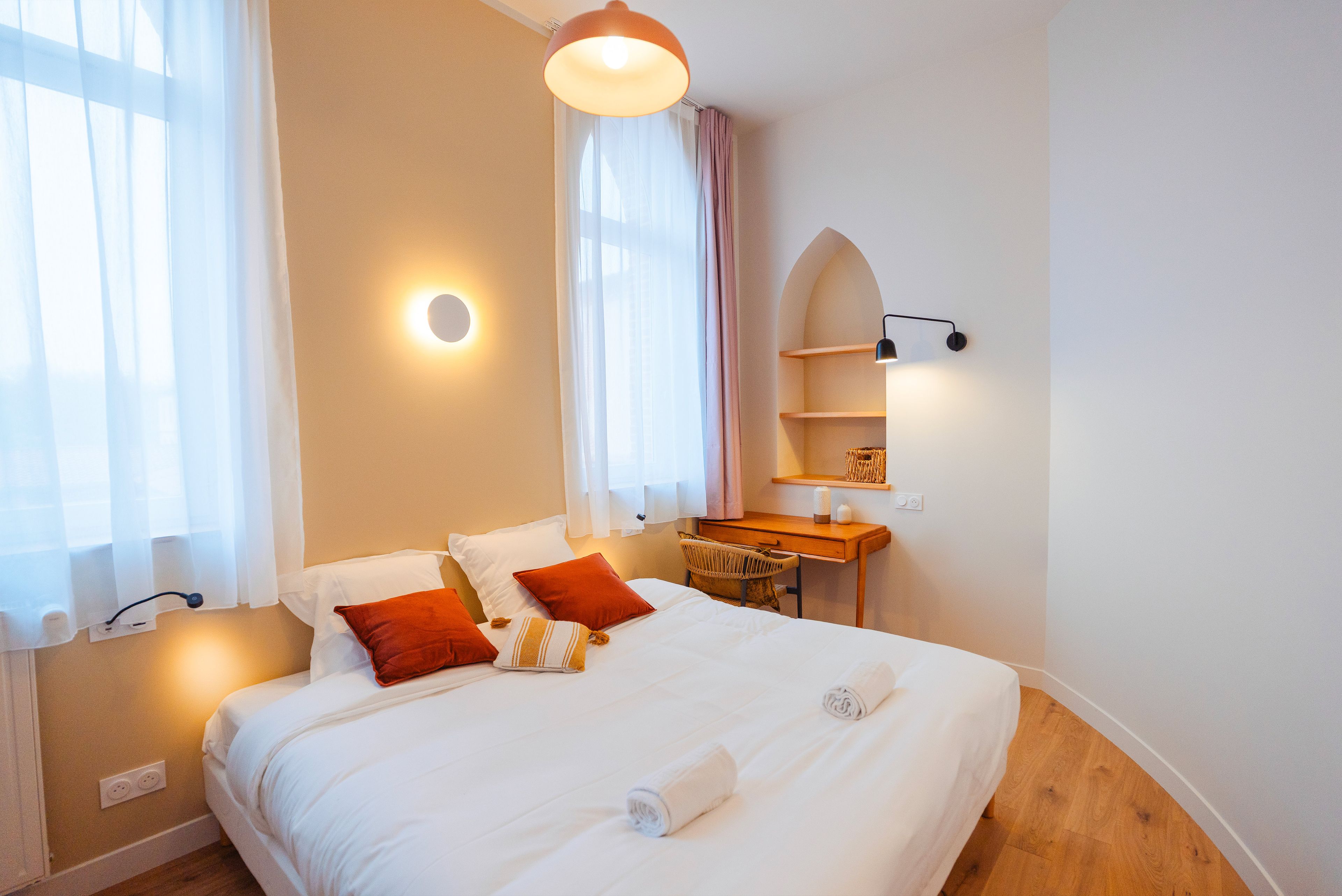 A hotel room with a large bed, a desk, and a chair. There is a window with sheer curtains and a wall-mounted shelf with a lamp.