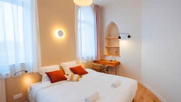 A hotel room with a large bed, a desk, and a chair. There is a window with sheer curtains and a wall-mounted shelf with a lamp.