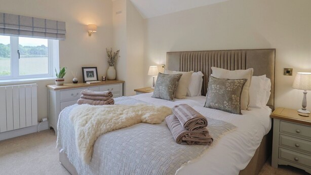 A bedroom with a large bed, a bedside table with a lamp, a window with a view, and a radiator. - The Fox Den (Yeovil)