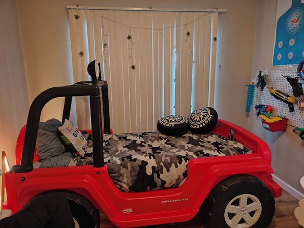 Ein Kinderspielbereich mit einem roten Spielzeug-Jeep Wrangler, Tarnfarben-Bettwäsche und einer Wandtafel mit Spielzeug.