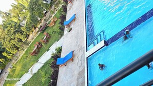 Een zwembad met een helderblauw wateroppervlak, omringd door een betegeld zonnedek met lounge fauteuils en een verzorgde tuin met diverse planten.