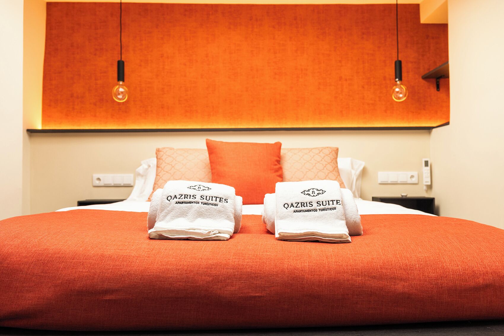 A hotel room with a bed, two pillows, a blanket, and two towels with 'Qazris Suites' branding.
