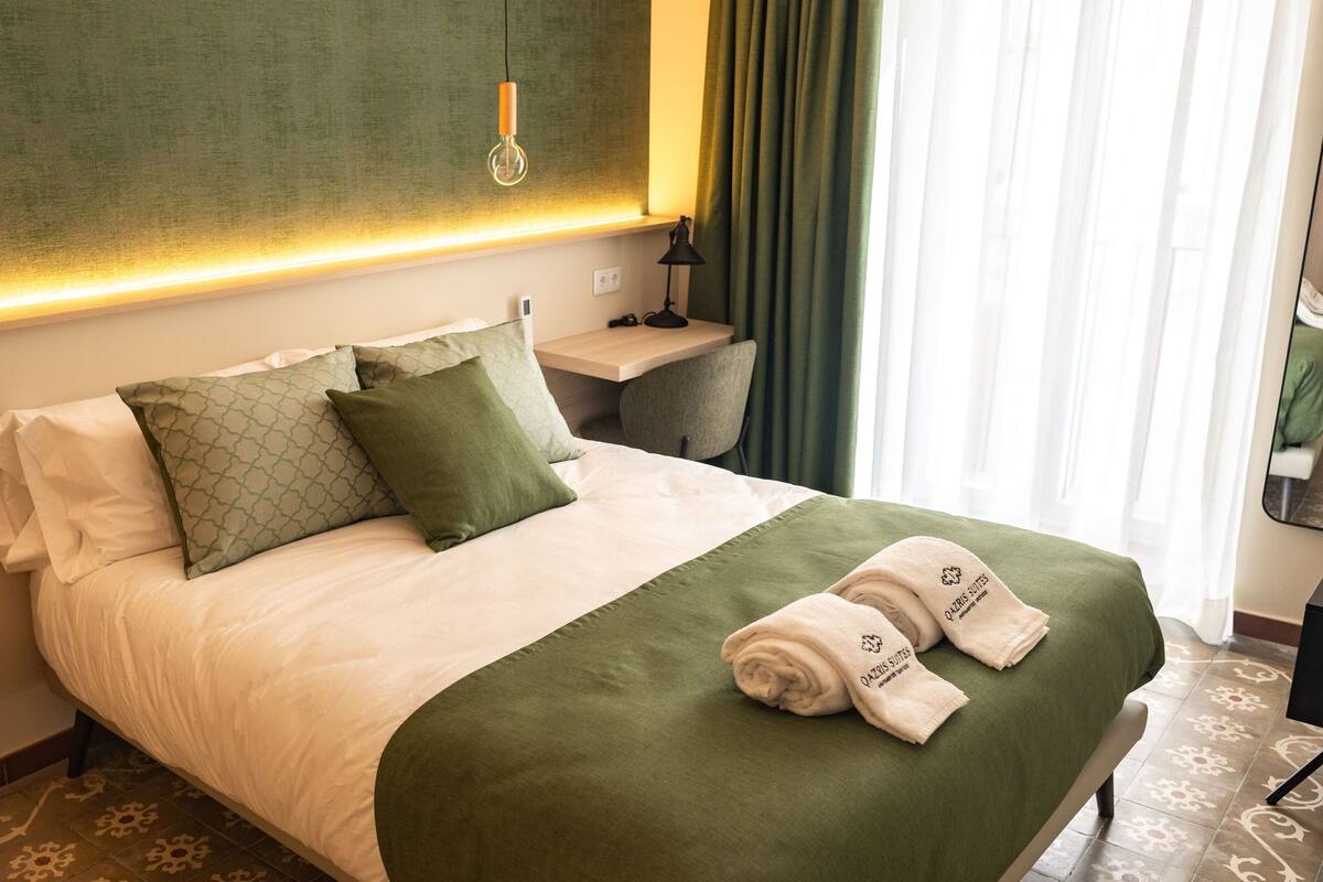 A hotel room with a neatly made bed, a bedside table, a lamp, and a view of the outside through the window.