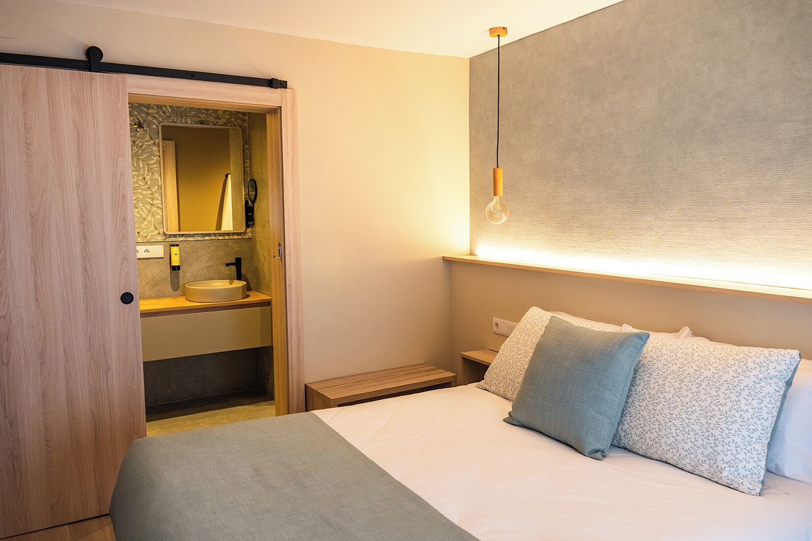 Habitación de hotel con cama, mesita de noche y armario empotrado con espejo y lavamanos.