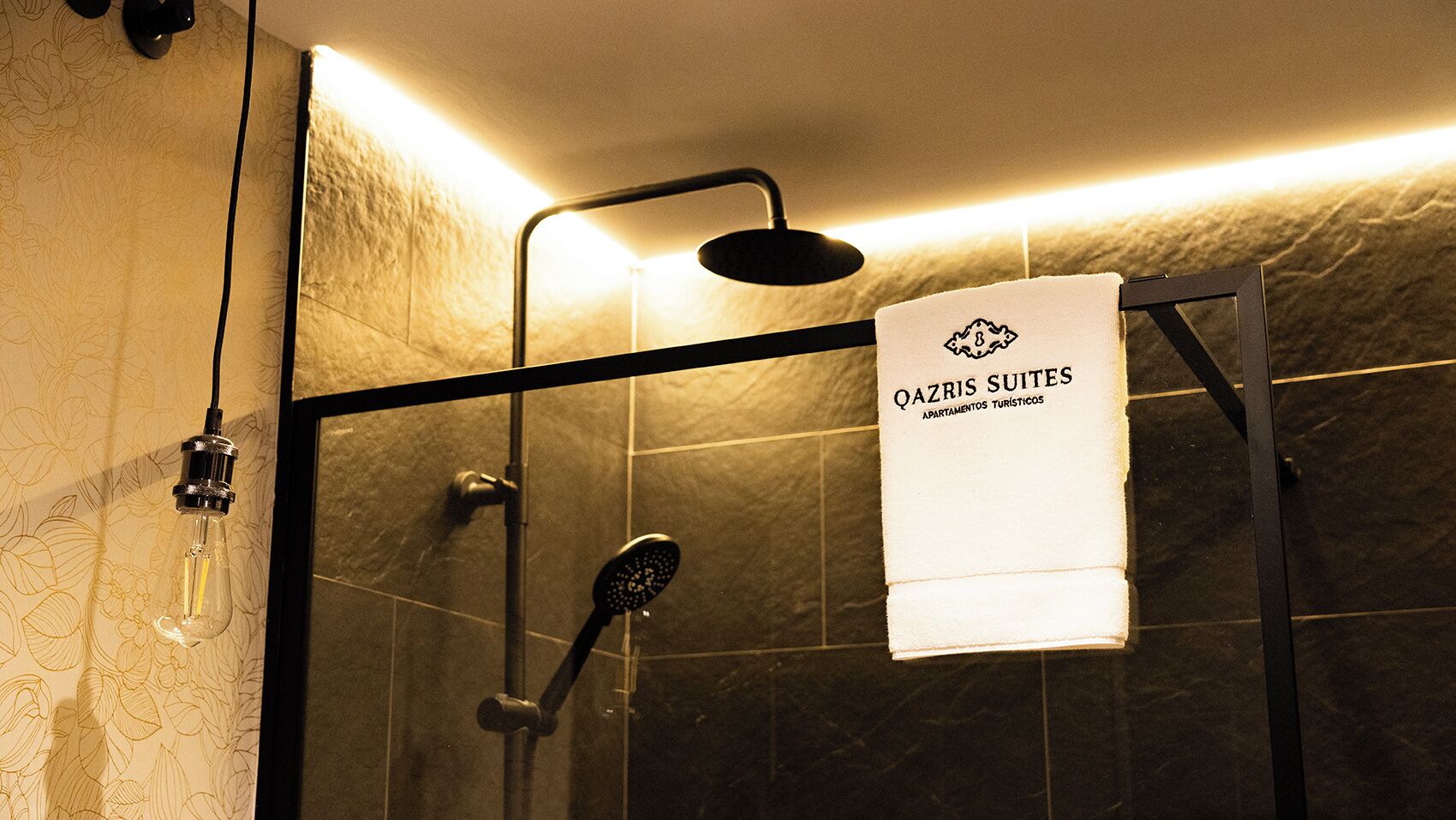 Ein Duschbereich mit Glastür, ein Handtuch, das an der Tür hängt, und ein Schild mit der Aufschrift 'QAZRIS SUITES APARTAMENTOS TURISTICOS'.
