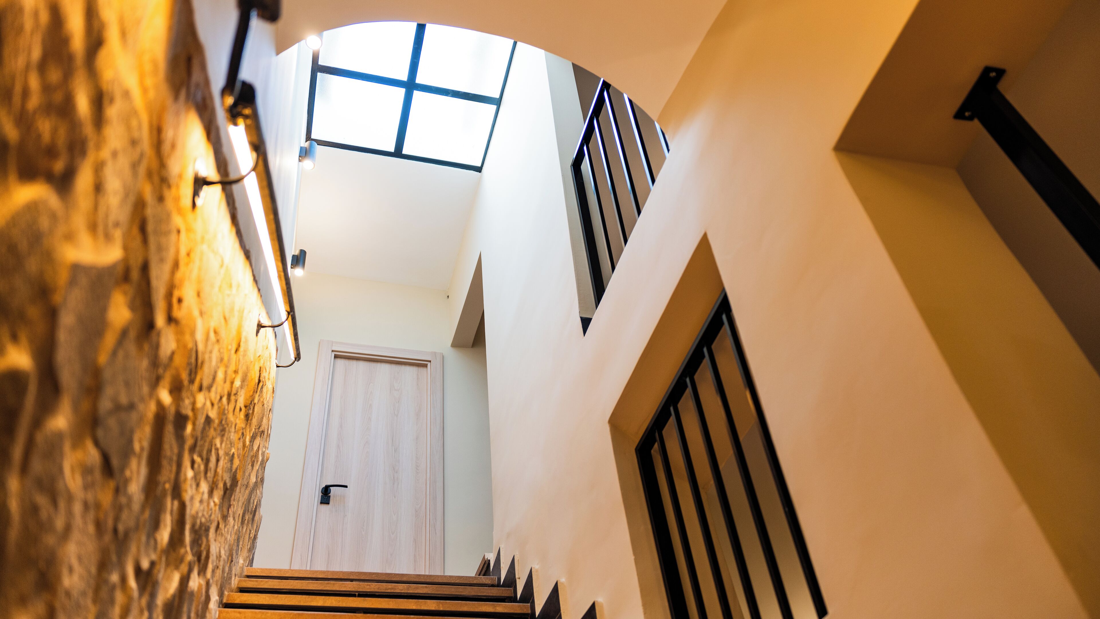 Un interior moderno con una escalera que conduce a un segundo piso, una puerta de madera y una vista del cielo a través de una ventana.