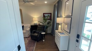 A hallway with a ceiling fan, a seating area with a couch and chairs, a small table with a lamp, and a window with curtains.