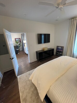 Un dormitorio con una cama, un televisor montado en la pared, un ventilador de techo y una puerta que da a otra habitación.