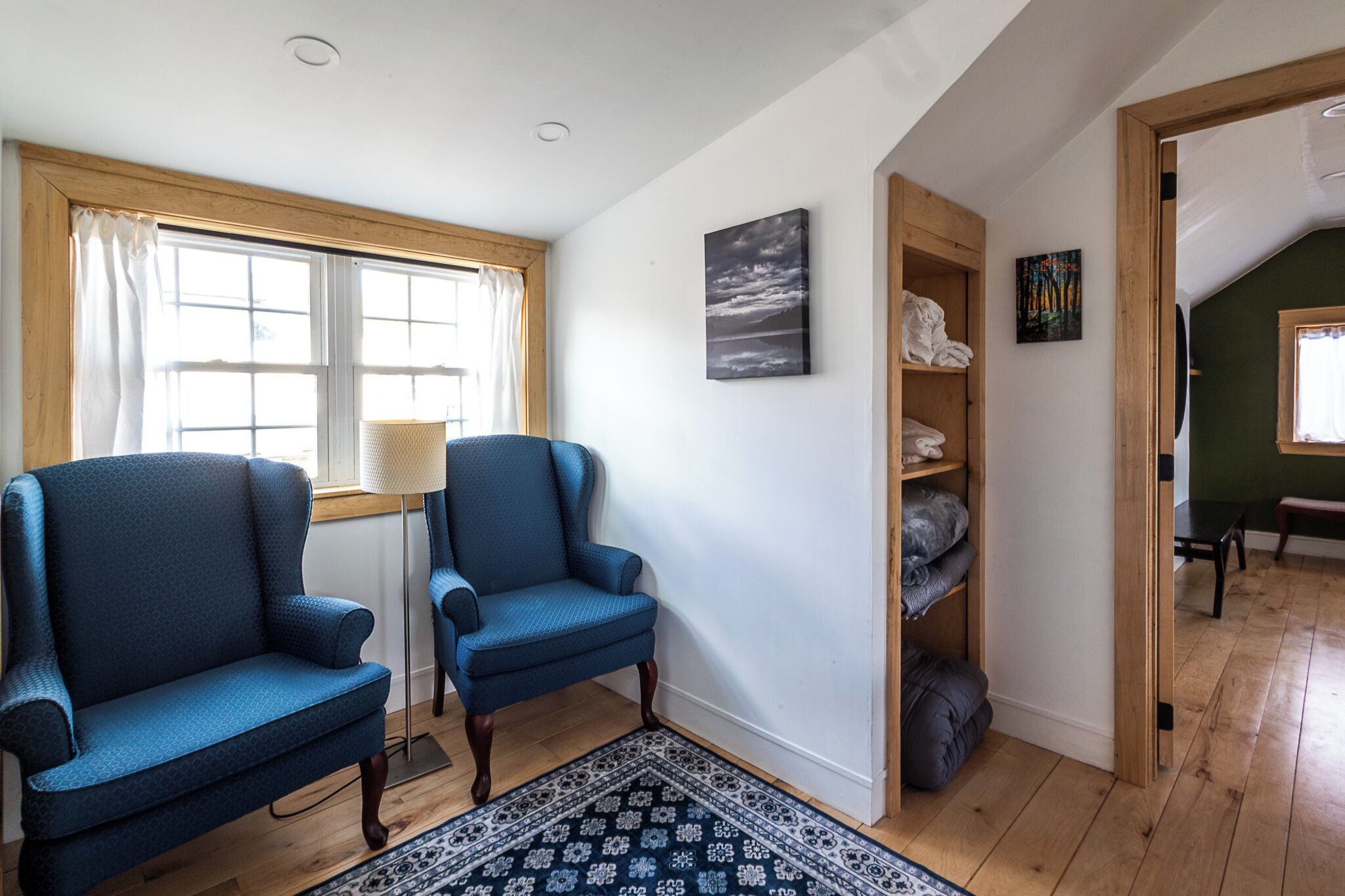 Una habitación con dos sillones azules, piso de madera, alfombra estampada, ventana con cortinas, estante con objetos doblados y un cuadro en la pared.