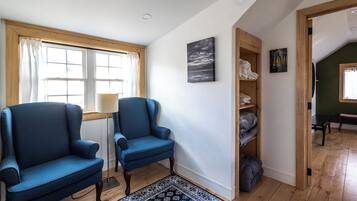 Una habitación con dos sillones azules, piso de madera, alfombra estampada, ventana con cortinas, estante con objetos doblados y un cuadro en la pared.