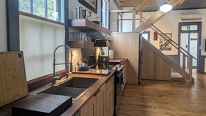 Une cuisine moderne avec des armoires en bois, un évier en acier inoxydable et une cuisinière intégrée. Une grande fenêtre offre une lumière naturelle et une vue sur l’extérieur. Un escalier en bois mène à l’étage supérieur.