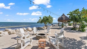 A beachside seating area with white Adirondack chairs, a stone fire pit, and a thatched-roof structure. - Freshly Renovated Coastal Stay • 2 Kings • Marina, Pool (Key Largo)