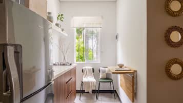 A compact kitchen with a refrigerator, a window with a view, a small table with a chair, and decorative wall mirrors.