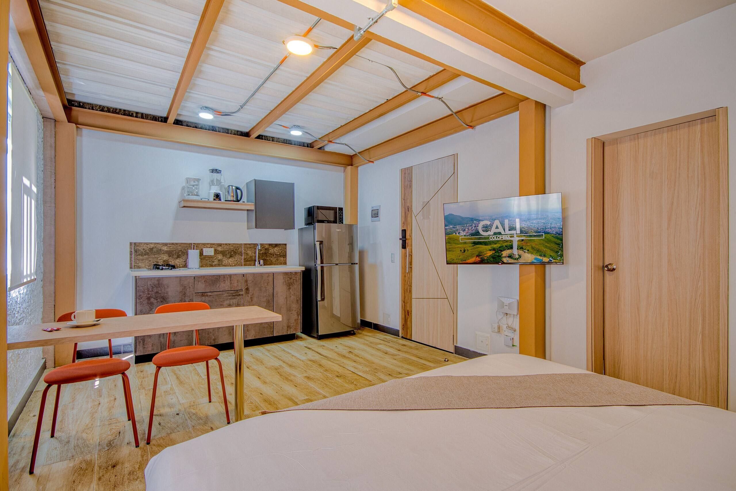 Una habitación compacta con cama, una mesa de comedor con sillas, una kitchenette y un televisor montado en la pared.