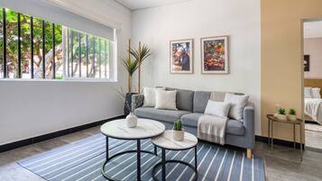 Ein modernes Wohnzimmer mit einem grauen Sofa, zwei runden Tischen und einem großen Fenster mit Blick auf Bäume.