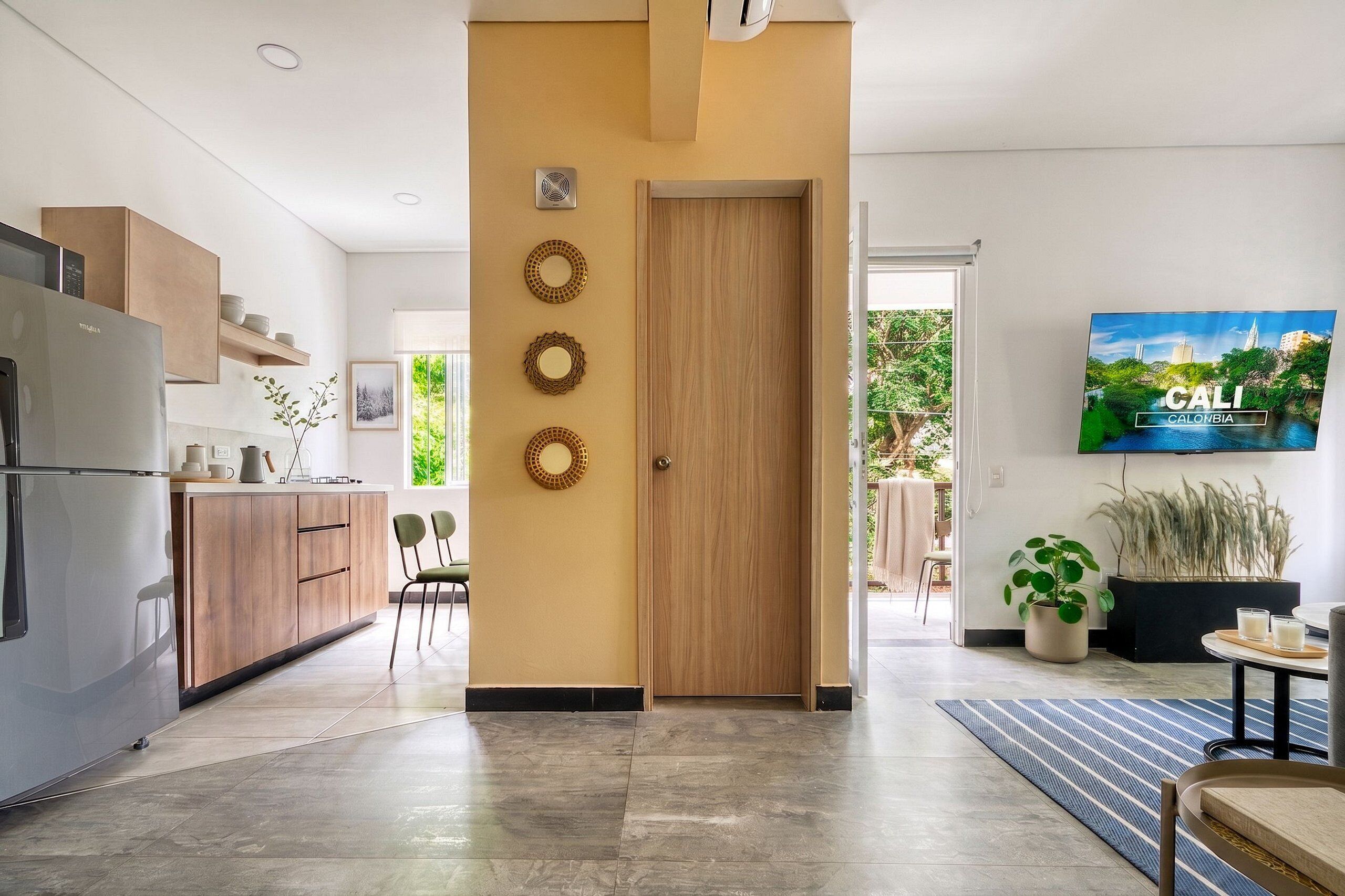 Una cocina moderna con puerta de madera, un comedor con sillas y un televisor mostrando un paisaje urbano.