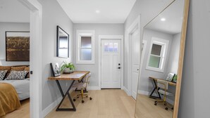 A modern, well-lit hallway with a wooden floor, a desk with a chair, a bed with a brown blanket, and a large mirror.