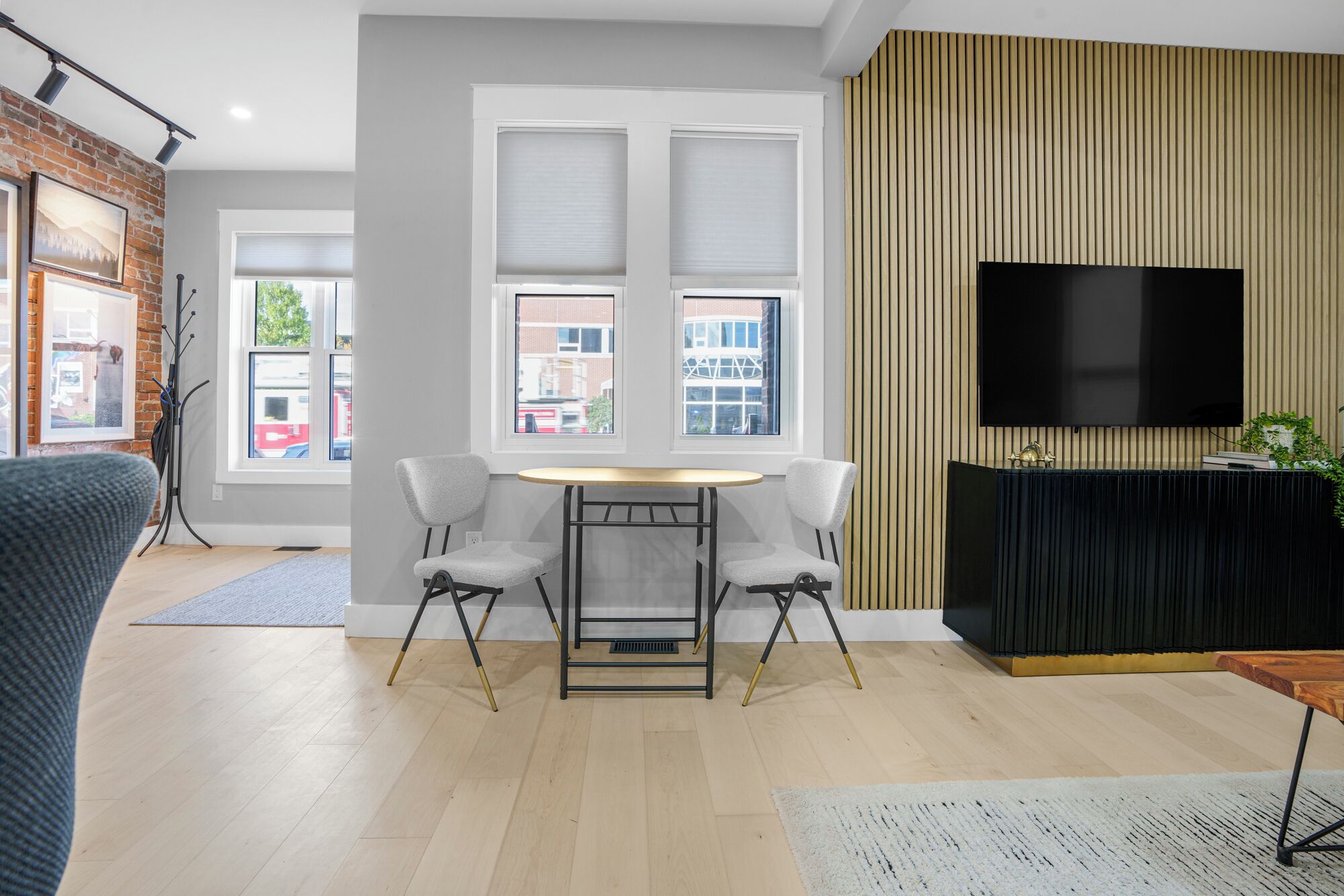 A modern living room with a wooden floor, a dining table with two chairs, a flat-screen TV mounted on the wall, and large windows with blinds.