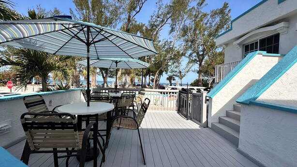An outdoor deck with tables and chairs, a striped umbrella, and a view of the beach. - Alamanda Villas (Holmes Beach)