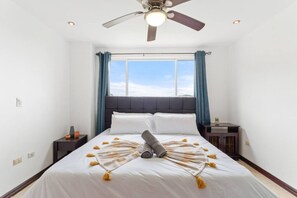 Un dormitorio con cama, ventilador de techo, ventana con cortinas y mesita de noche con lámpara.