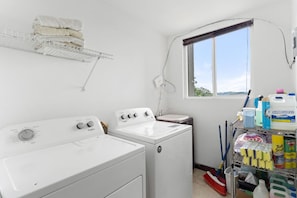 Un cuarto de lavandería con dos lavadoras y secadoras Whirlpool blancas, un estante con toallas dobladas, un ventanal y un estante para productos de limpieza.