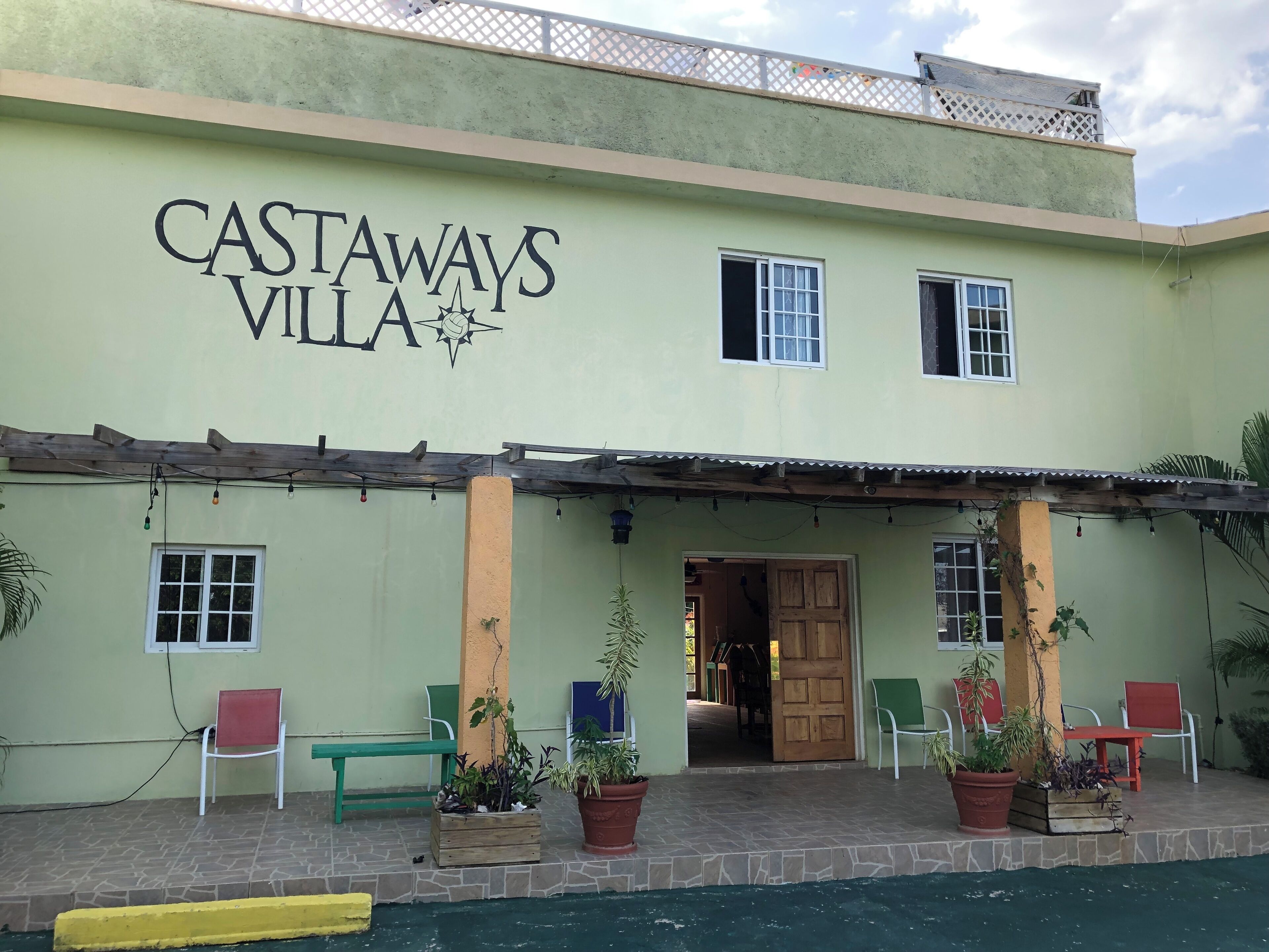 Een gebouw met het bord 'CASTAWAYS VILLA' en een overdekt entreegebied met stoelen en plantenpotten.