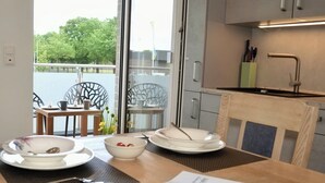 A dining area with a table set for two, a view of the outdoors, and a modern kitchen with a sink and cabinets. - KomfortPlus New Construction - Apartment with a sunny balcony and free parking (Bad Zwischenahn)