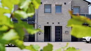 A multi-story building with a brick facade, two black doors, and a white car parked in front. - KomfortPlus New Construction - Apartment with a sunny balcony and free parking (Bad Zwischenahn)
