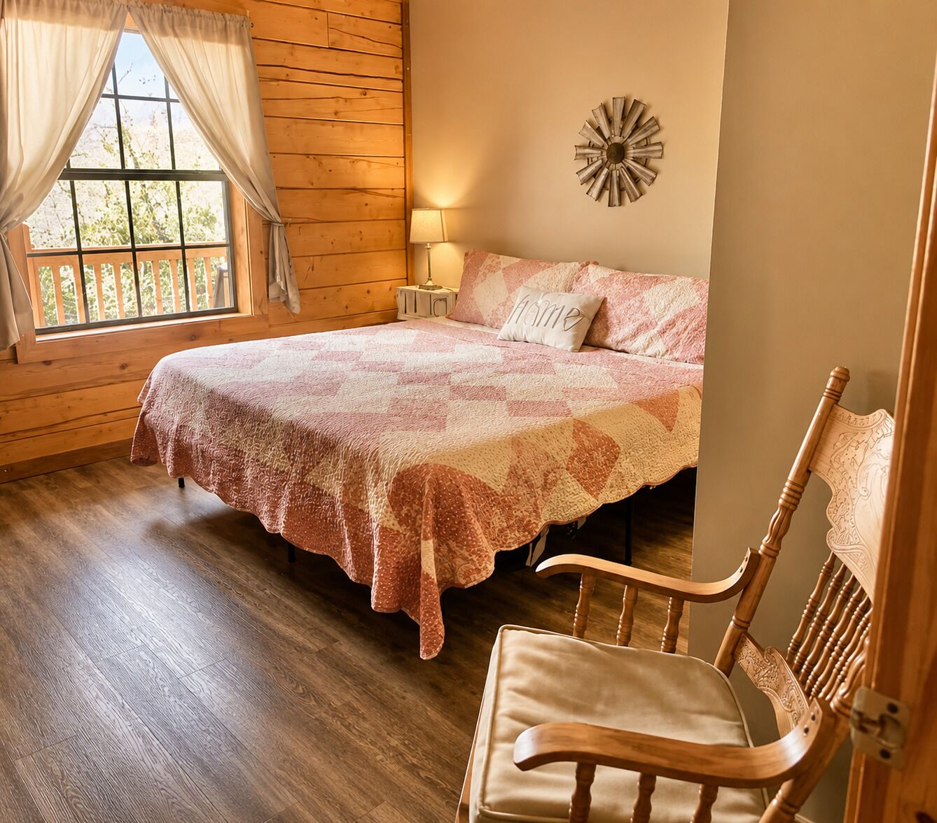 Une chambre avec un plancher en bois, un lit recouvert d’une couvre-lit à motifs, une table de chevet avec une lampe et une fenêtre avec des rideaux.