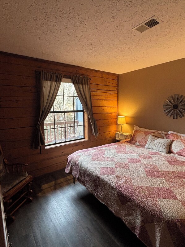 Un dormitorio acogedor con suelo de madera, una cama grande con colcha rosa y blanca, una ventana con cortinas y una lámpara fijada a la pared.
