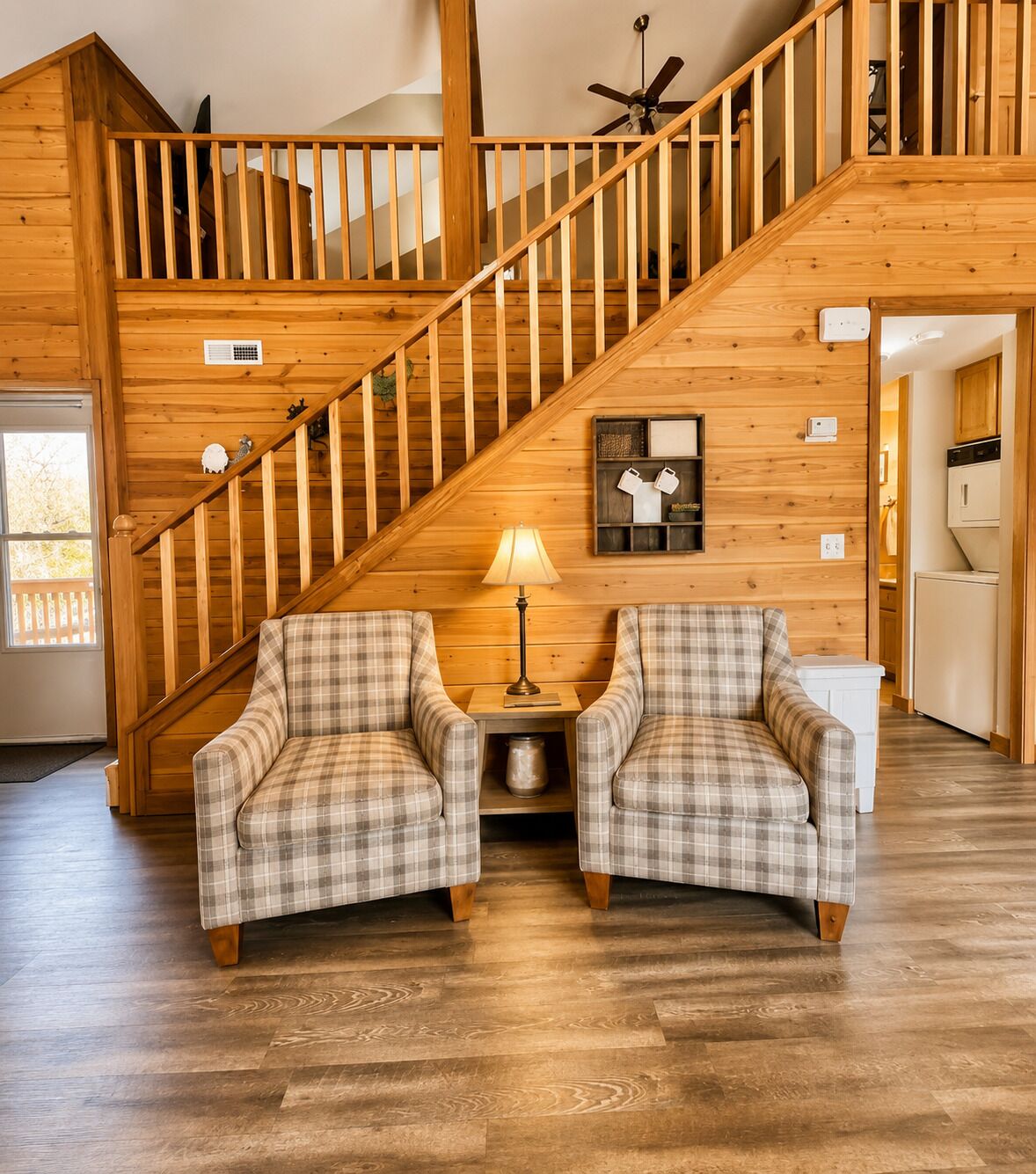 A wooden staircase leading to a loft area with a ceiling fan, a lamp on a side table, and two plaid armchairs facing each other.