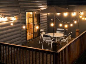 Una terraza de madera con una mesa redonda y sillas, luces colgantes y un paraguas grande.